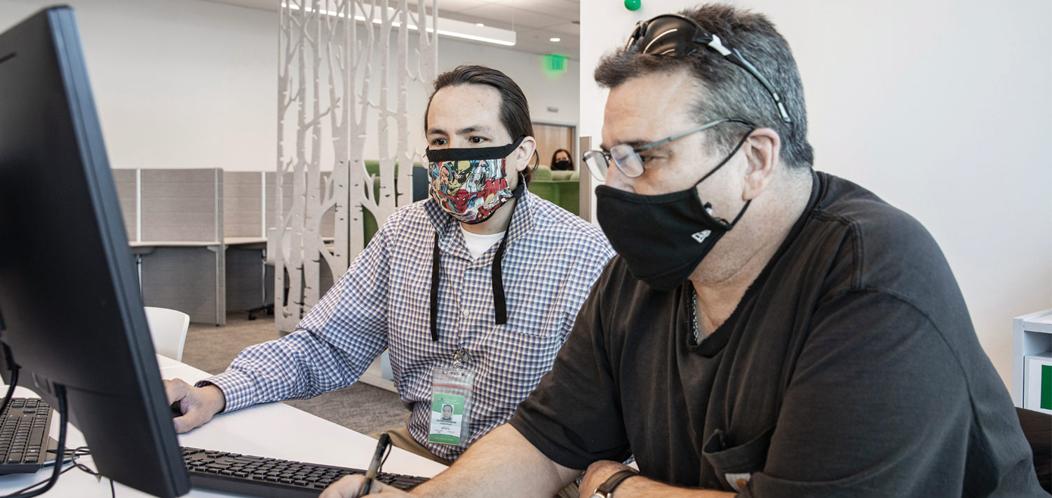 two people wearing masks sit a computer together
