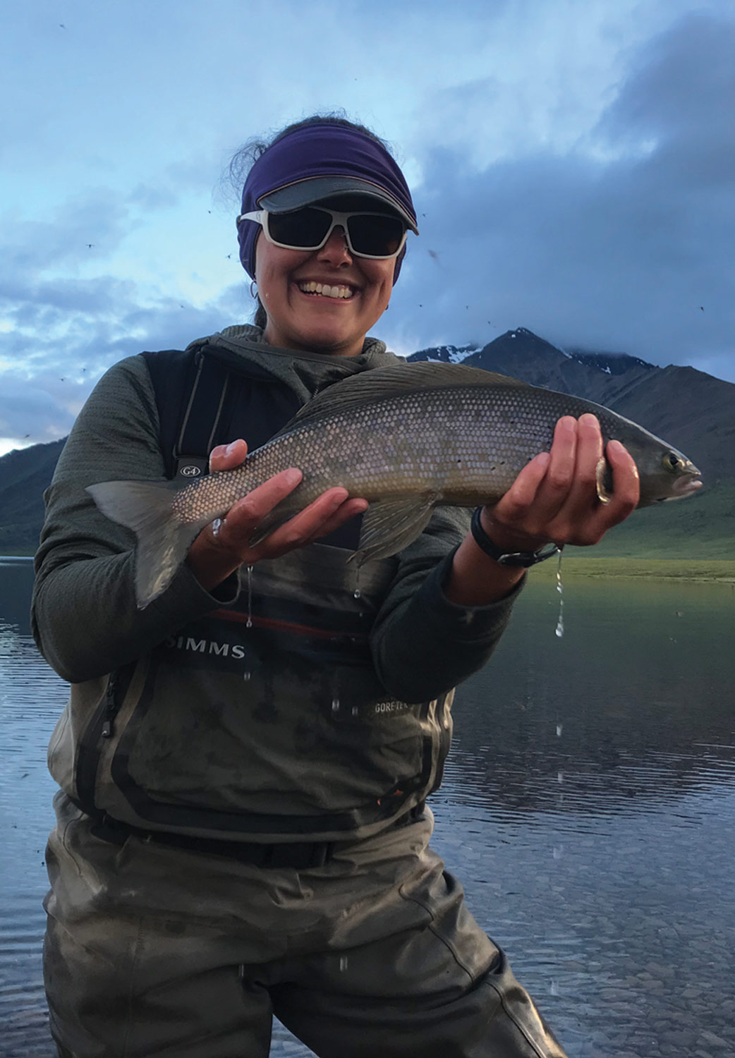 a woman smiles holding an Arctic grayling while standing in water