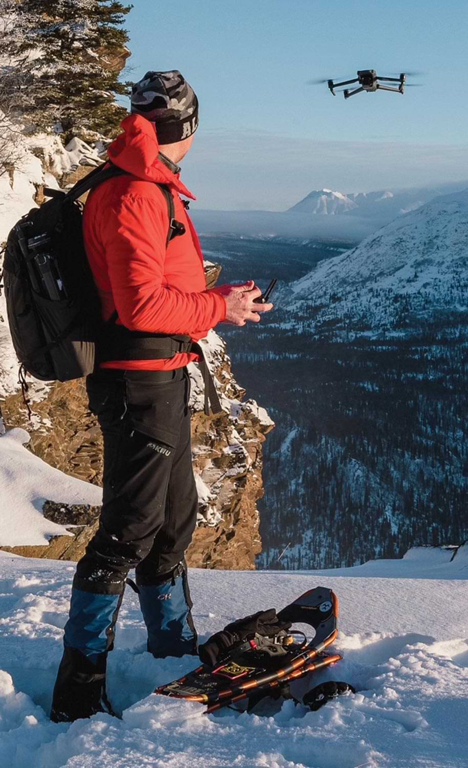 a man dressed in snowboarding gear operates a drone from a snowy mountain ledge