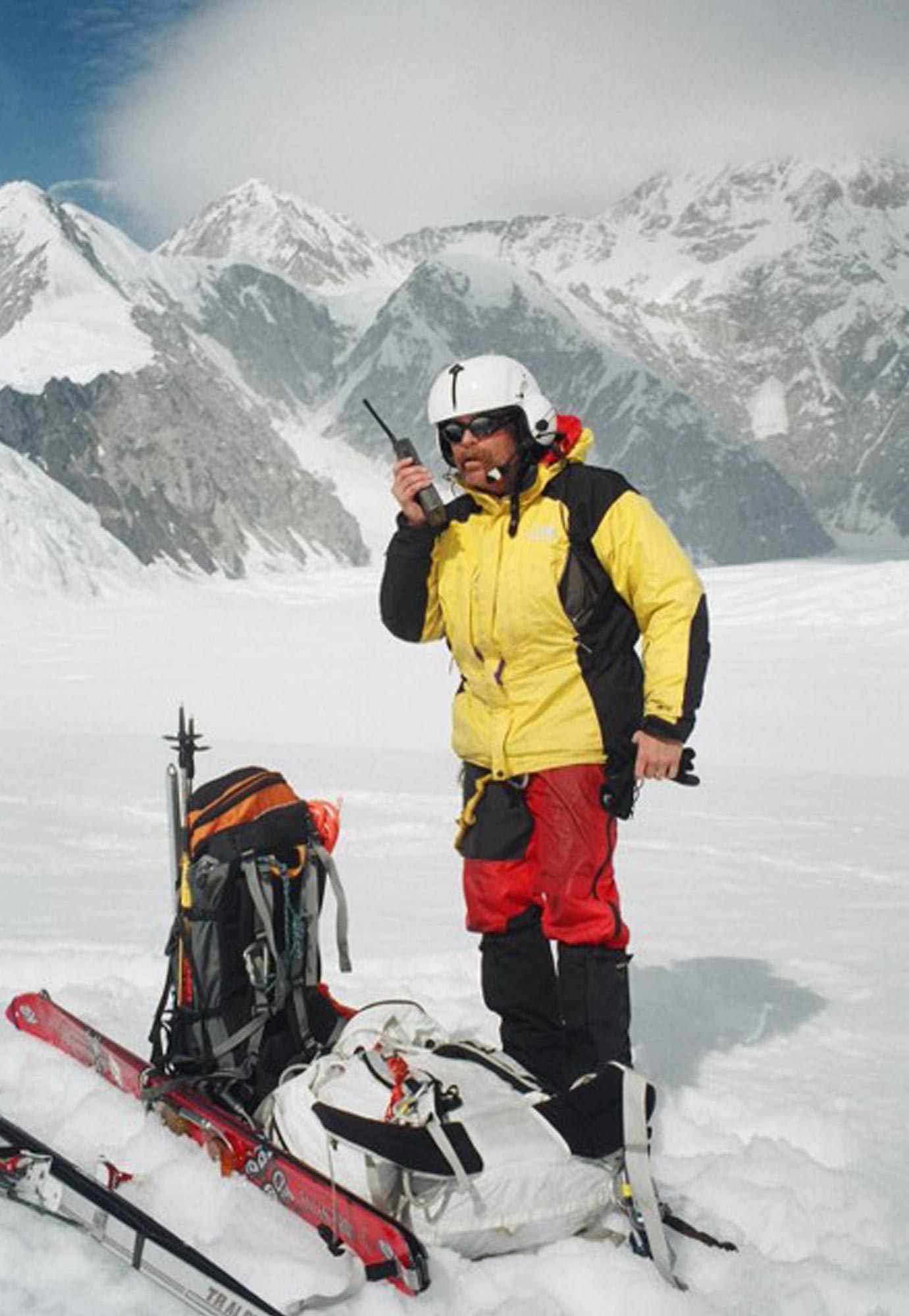 a man wears ski gear and speaks into a walkie-talkie while standing on a snow cover mountain plateau