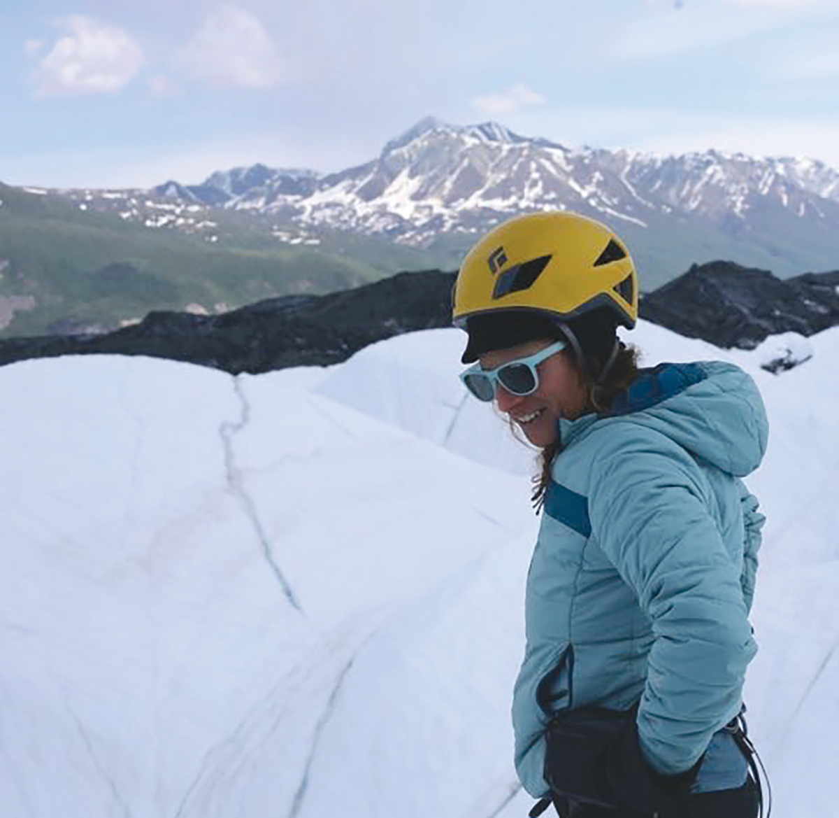A portrait photograph of a woman in a blue puffy jacket with blue sunglasses and a yellow helmet is smiling looking away from the landscape backdrop of a snowy mountain