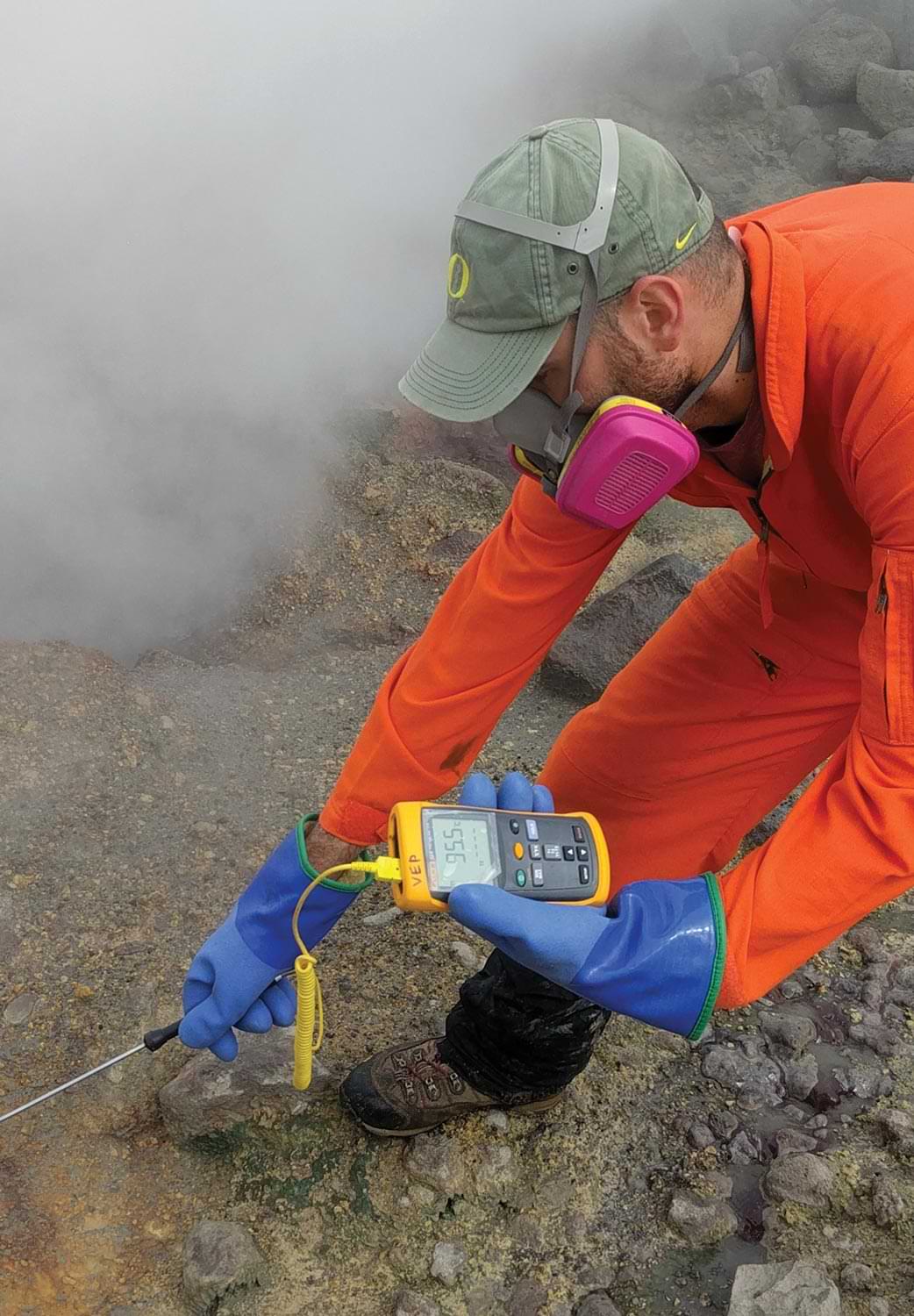 geochemist Allan Lerner of the US Geological Survey wears a ventilation mask, blue gloves and a bright yellow jumpsuits while using a device to measure the temperature of a bubbling pool in the upper Glacier Valley south of Makushin Volcano