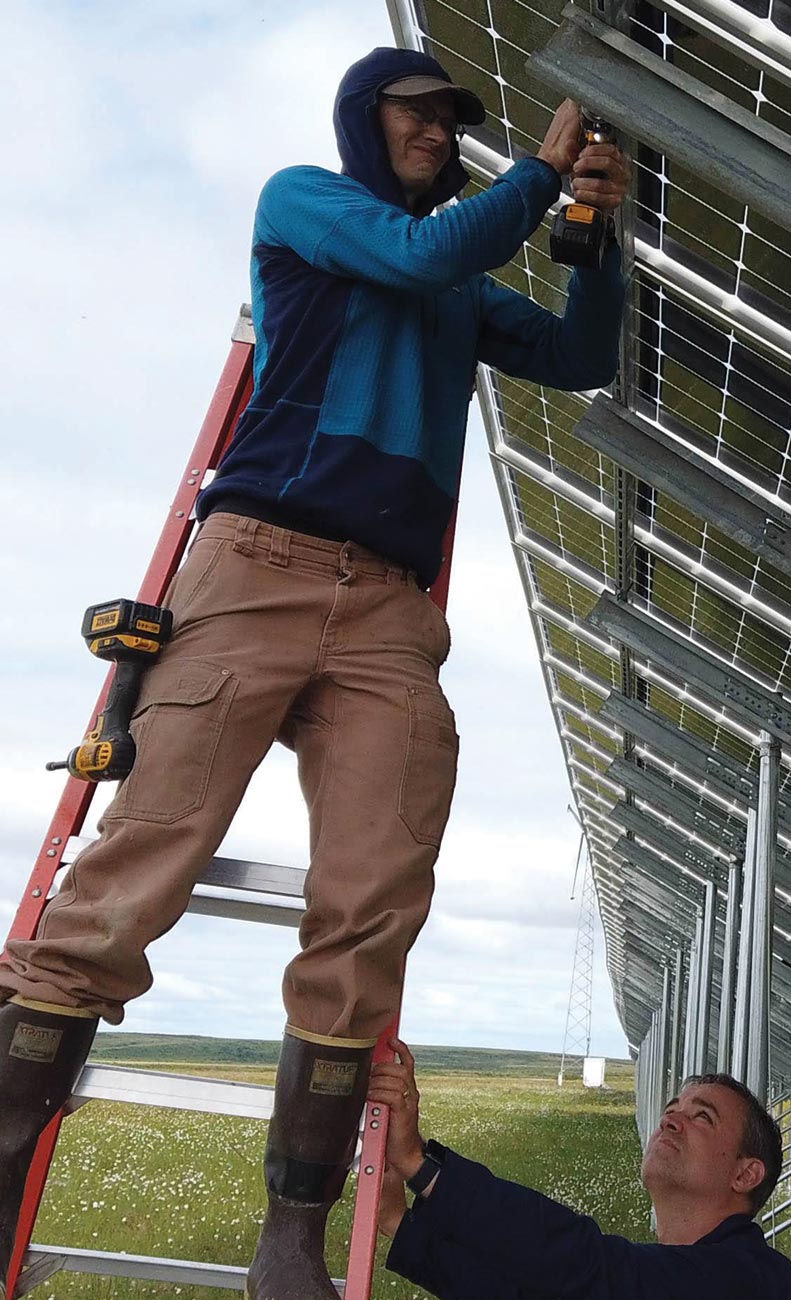 Alaska Center for Energy and Power’s Chris Pike stands on a ladder and uses a drill to install a monitoring camera on a solar panel in Kotzebue