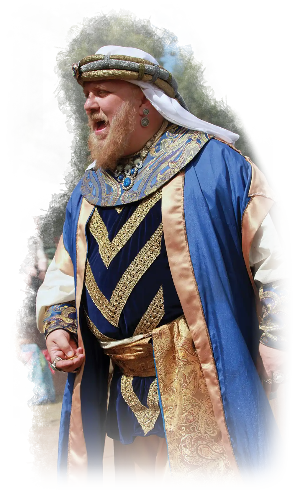 A man is dressed in a Middle Eastern-style embroidery fabric ornate costume with a multi-colored silk hat (white, dark gold/bronze, and faded silver) plus several jewel rings on his fingers as his mouth is slightly open indicating a yelling facial expression