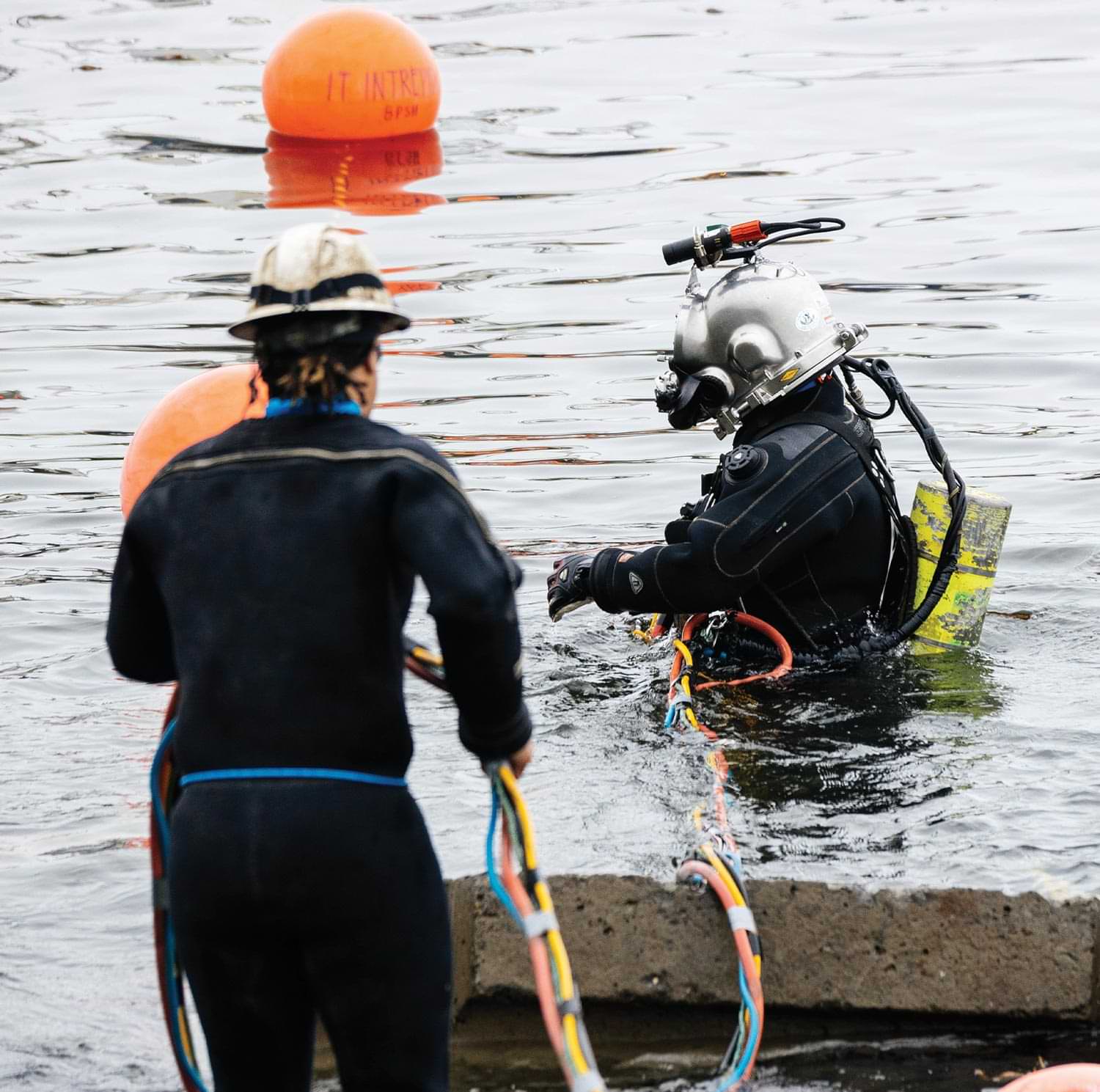 a diver in water wears special gear as a person in a wet suit holds thick taped fiber cables from a rocky shore during the AU-Aleutians Fiber Project