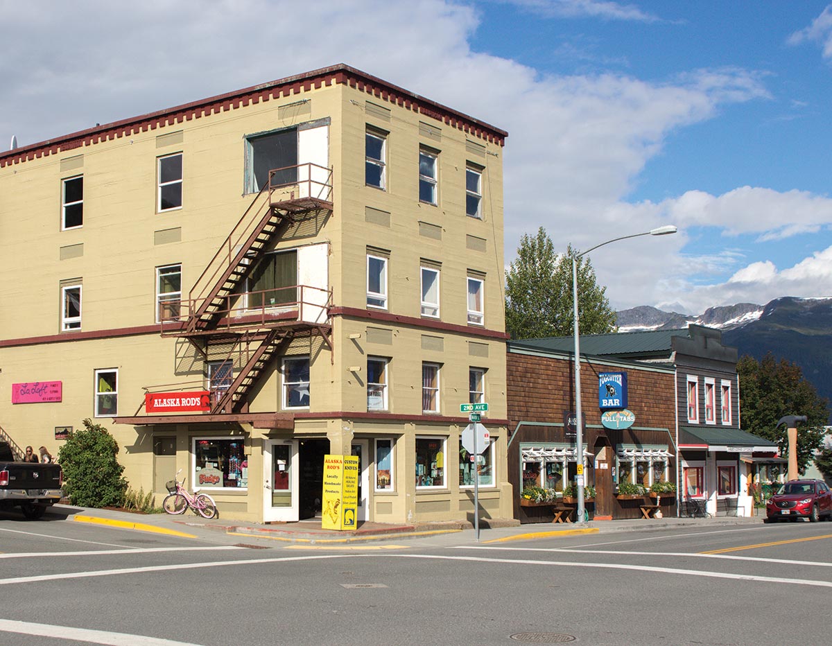 Angle corner street photograph perspective of a light tan/dark brown colored building "Alaska Rod's" in Haines, AK during the day with some clouds in the sky