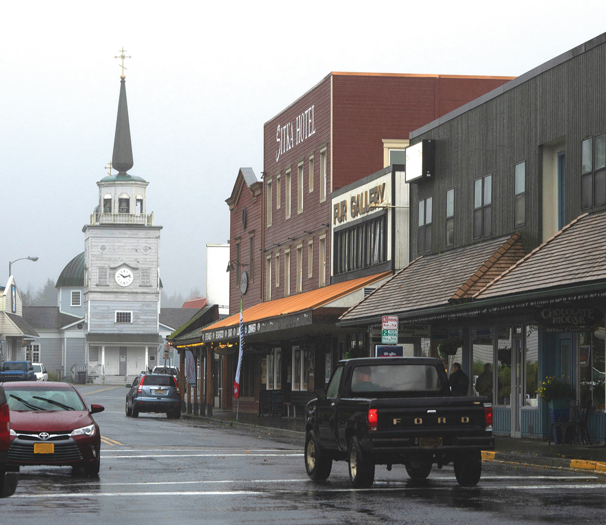 Portrait street corner angle photograph perspective of the Sitka Hotel & Sitka Fur Gallery buildings and many other businesses nearby with people driving their vehicles on the street on a gloomy, foggy day