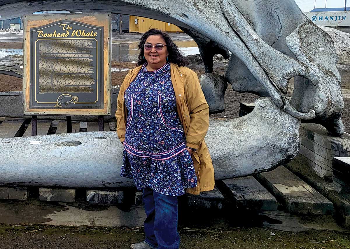 Landscape photograph of Frieda Nageak smiling as she poses next to The Bowhead Whale sculpture display; She loves showing visitors around Iḷisaġvik College, where she works as external affairs coordinator. Her idea for a souvenir shop earned her a spot in the 2023 cohort of North Slope Marketplace Business Boot Camp.