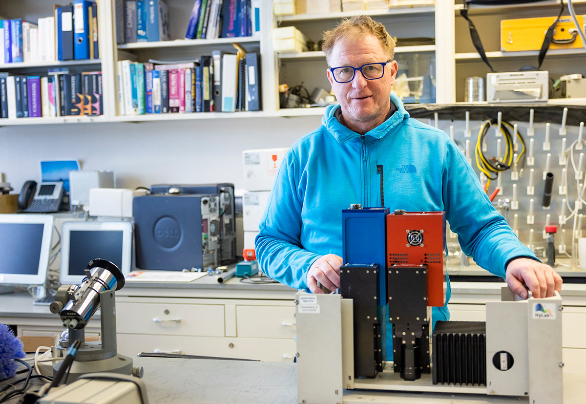 A hyperspectral camera system is usually mounted on a Aviat Husky airplane to collect surface data to find minerals. Martin Steufer, director of UAF Geophysical Institute's Hyperspectral Imaging Laboratory (pictured standing in front of the lab equipment machine in a lab setting environment), says hyperspectral imaging allows mining companies to identify potential locations remotely in the early development stages.