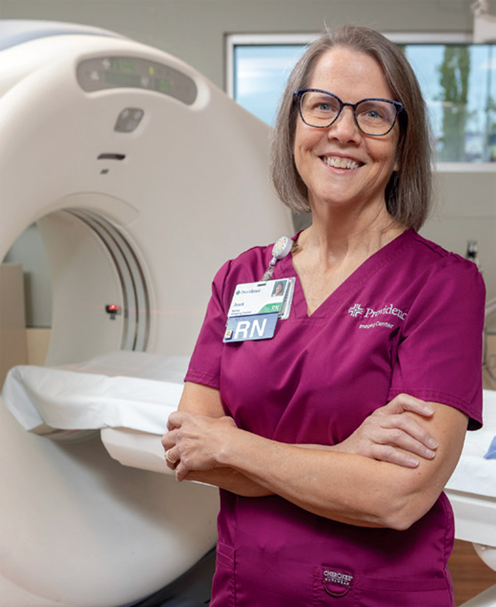portrait image of nurse Jean Dore wearing magenta scrubs emroidered with the Providence logo and standing in front of a CT scanner