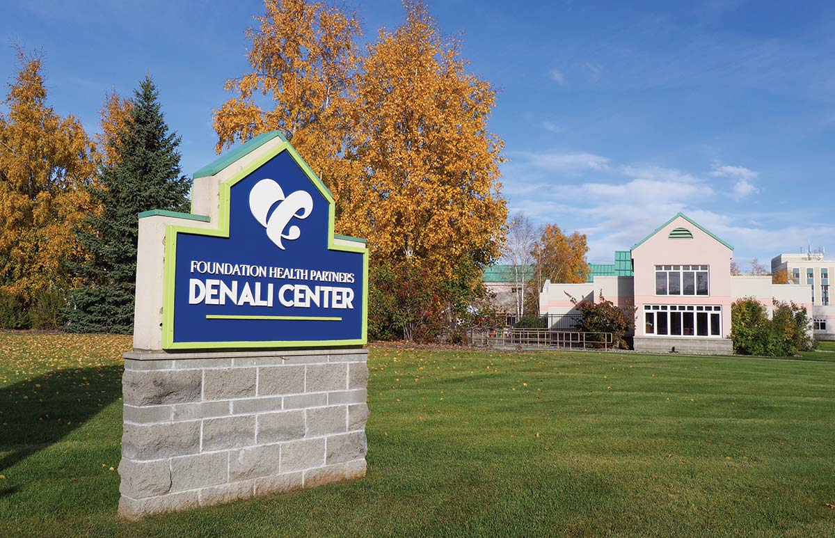 Landscape photograph of the blue/white Foundation Health Partners Denali Center logo brick wall front entrance sign situated on a grassy lawn area while in the background are several interconnected buildings