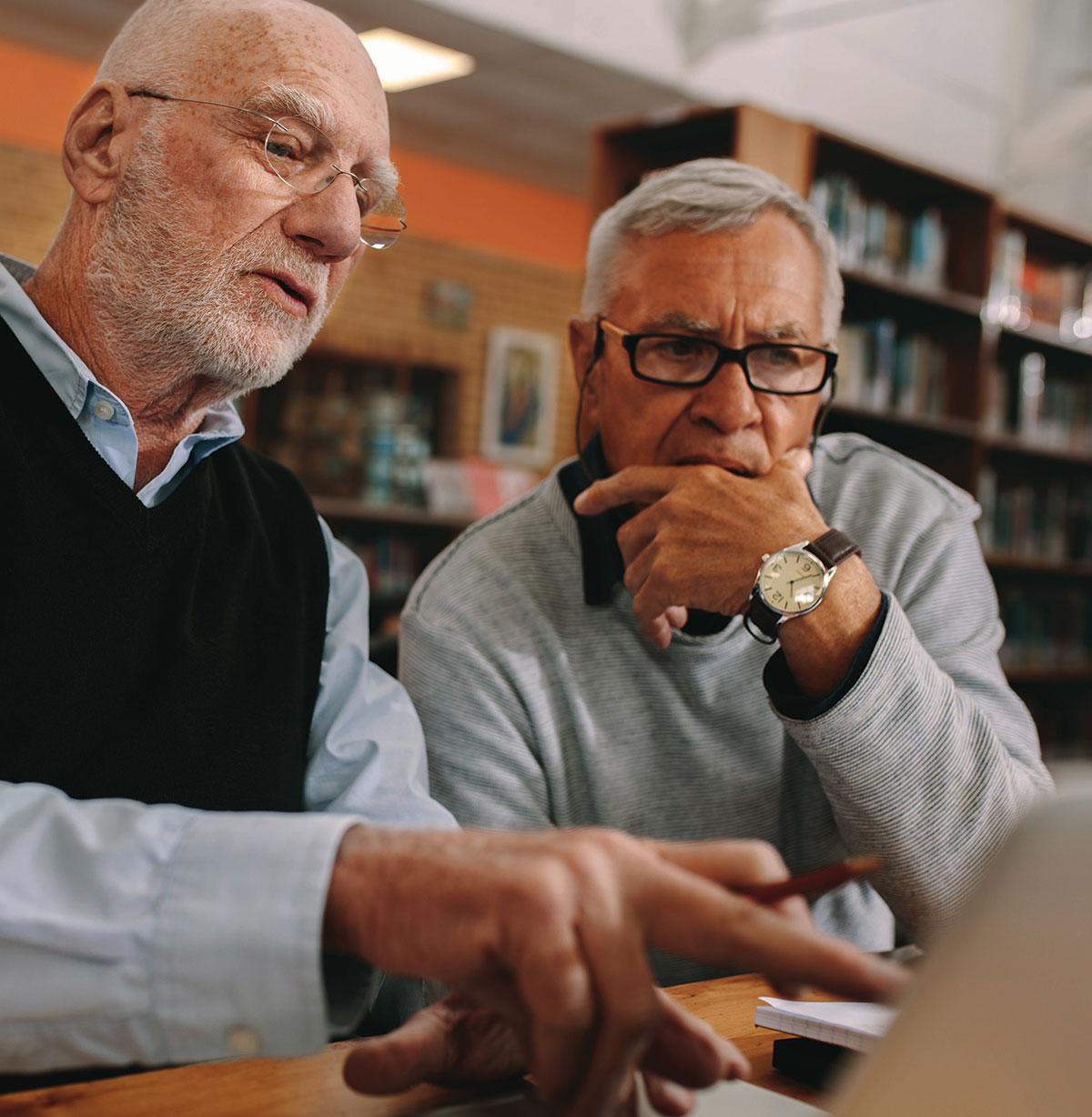 Close-up portrait photograph perspective of two older elderly men in see through prescription glasses glancing toward a laptop screen as one of the men has a pencil and is pointing with his finger at the laptop screen as they are in a public library setting