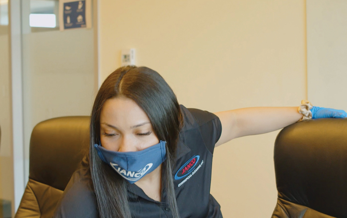 Janitorial woman worker from Janco Commerical Cleaning in a black company polo shirt inside an office area glancing down or cleaning likely at a table surface area holding onto some office chairs as she is also wearing blue medical gloves and a Janco company face mask