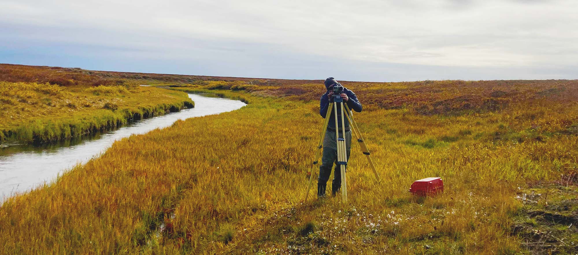 Man looking through a camera on yell tripod, in tundra landscape with a stream next to him