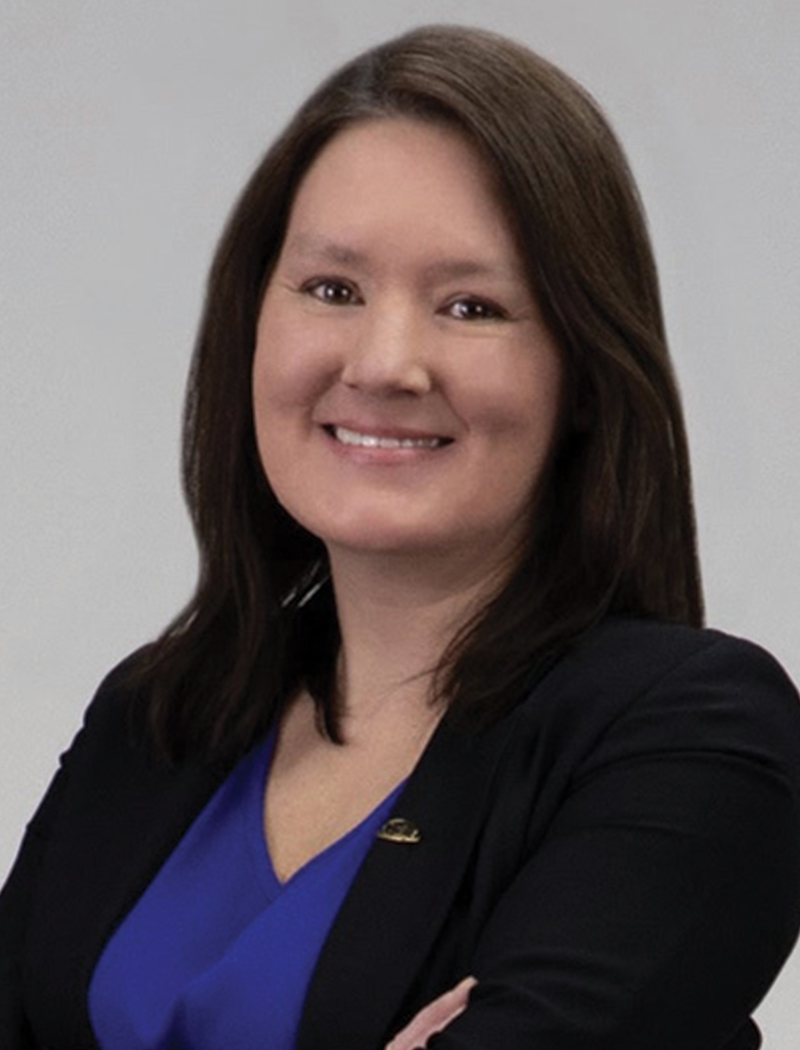 Headshot of a woman with dark hair, wearing a black blazer and blue shirt (Katherine Carlton)