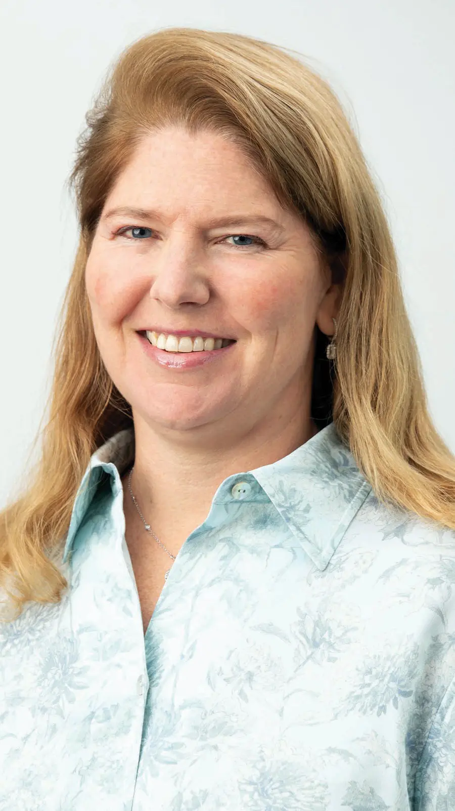 Portrait headshot photograph of Christine Resler smiling in a faded light aqua marine blue/white floral design pattern button-up shirt