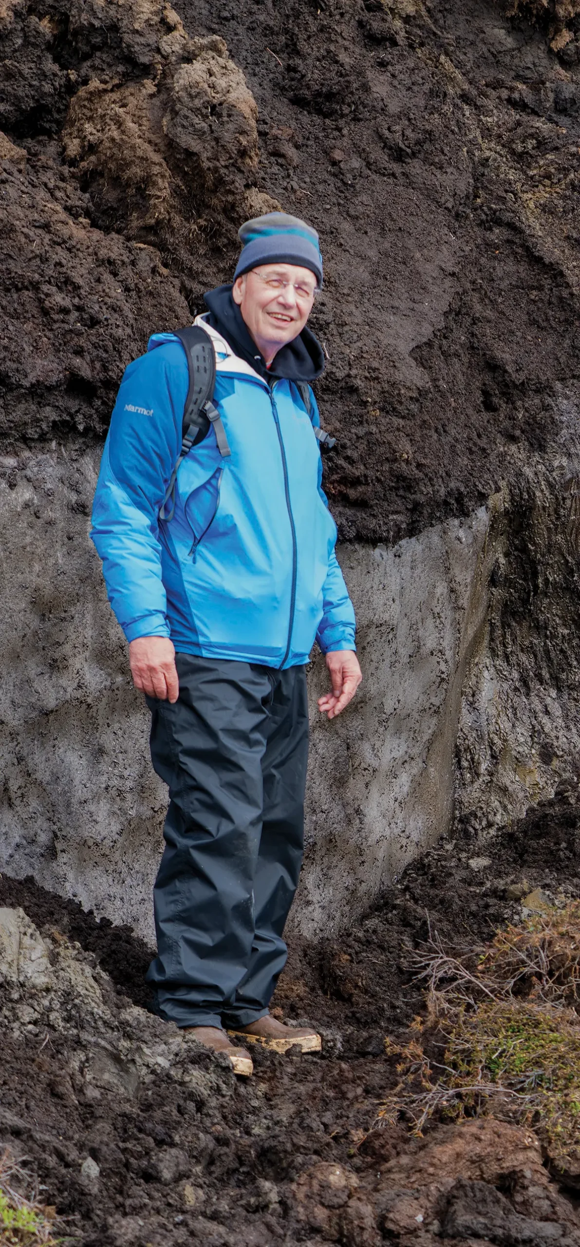 Portrait photograph of Billy Connor smiling in hiking attire wearing a multi-colored beanie (color shades of dark blue and dark grey), light blue jacket, black sweatpants, dark brown boots, and has a backpack on his shoulders standing outside somewhere near a cliff wall