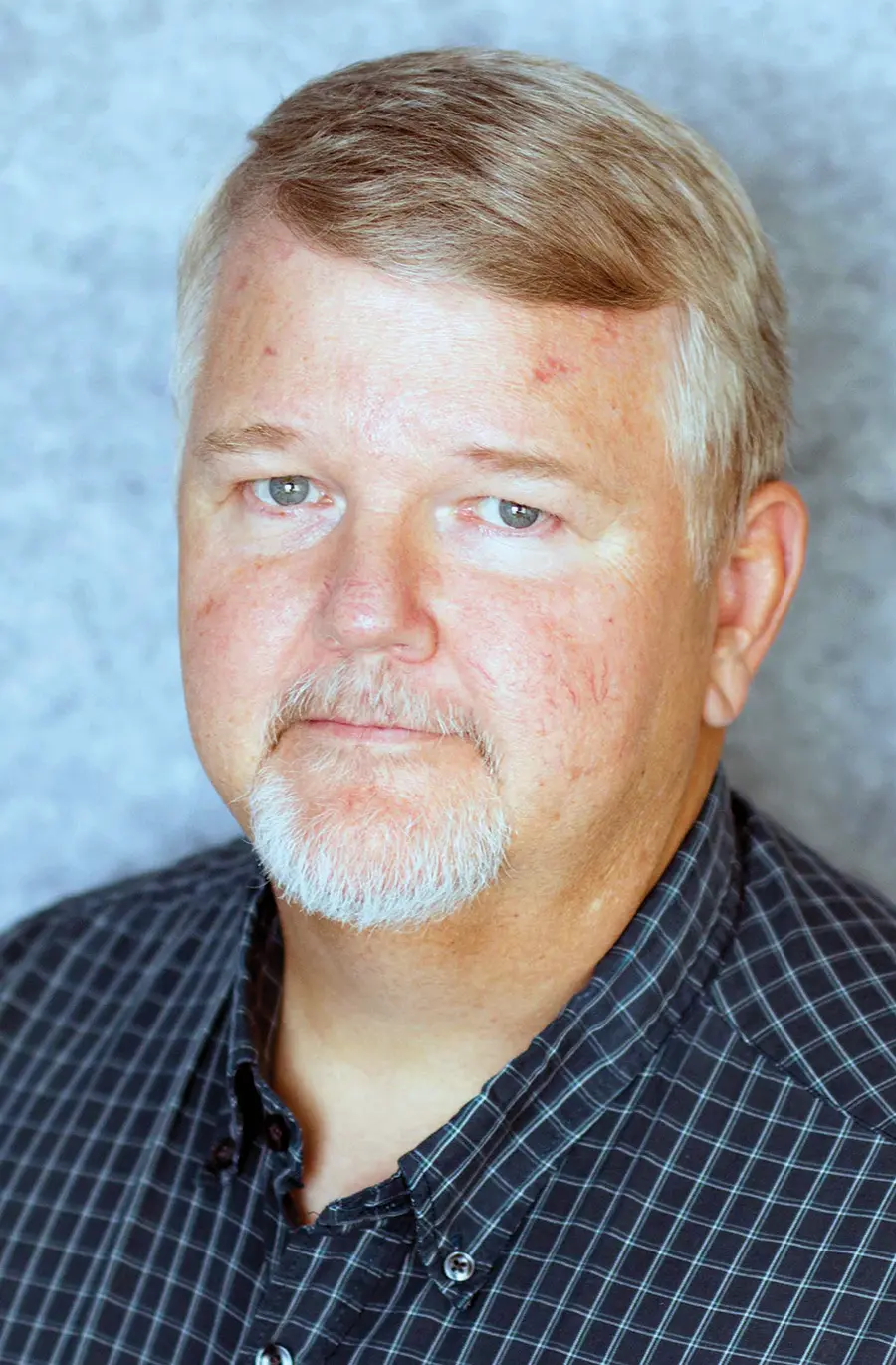 Portrait headshot photograph of Tim Collett grinning in a dark navy blue/turquoise line striped pattern plaid button-up shirt