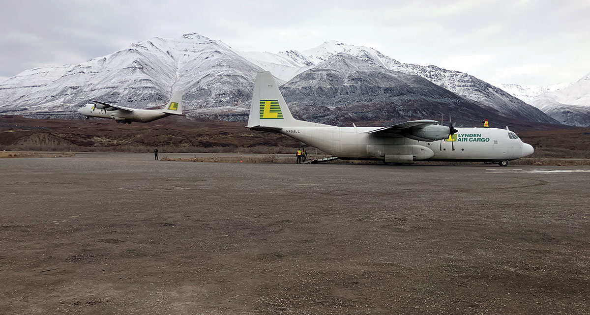 Landscape photograph of two grey Lockheed C-130 Hercules aircraft planes with the Lynden Air Cargo brand logo proudly displayed on them as one of the planes is flying in the other direction and the other is grounded on some dirt nearby some snowy mountain hillsides while there are a couple individuals in flight construction attire standing around on ground on a gloomy day