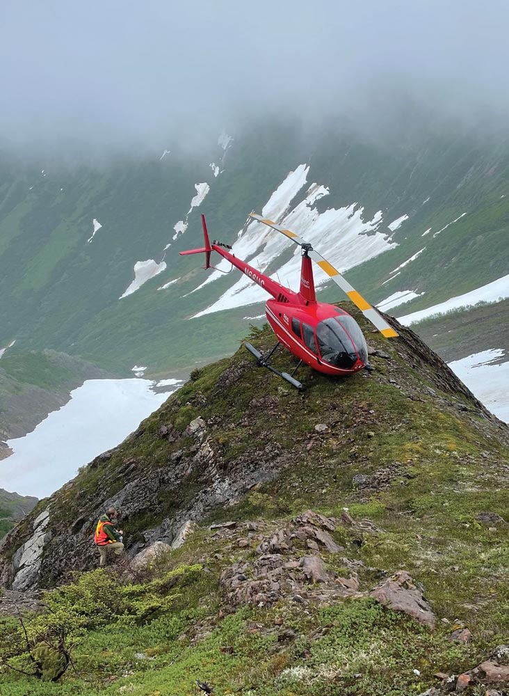 distant high angle view of a red helicopter resting at the top of a peak on the edge of a valley, a man in a safety vest surveys the ground on the slope just below the helicopter