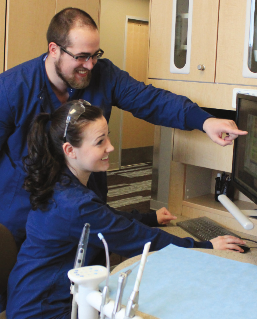 male dental assistant pointing to computer screen while a female assistant sits smiling with her hand on computer mouse