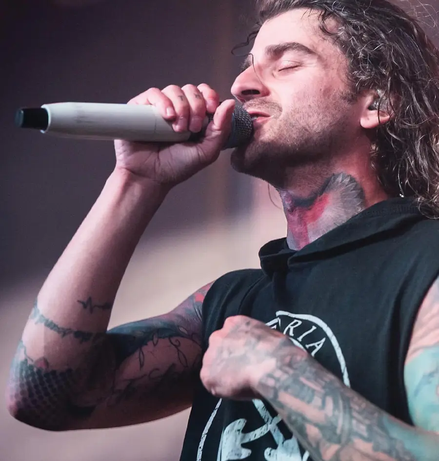 close view of a man with long wavy hair and many tattoos holding a microphone with his eyes closed while performing on a stage