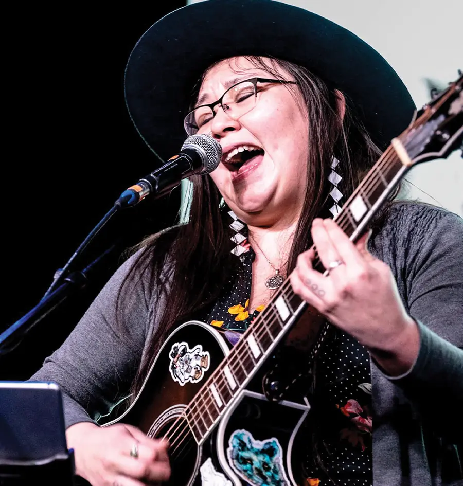 a female musician wears a wide brim hat and sings into a microphone as she plays an acoustic guitar