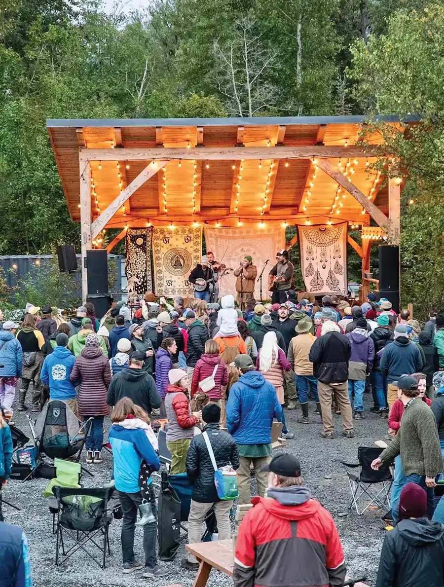 a crowd stands in front of a band playing on a small wooden stage at a small scale outdoor venue