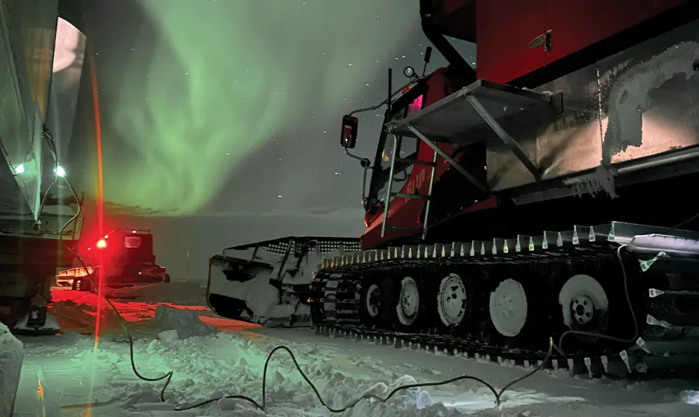 a large snowcat plowing as a green aurora borealis looms over the CWAT route