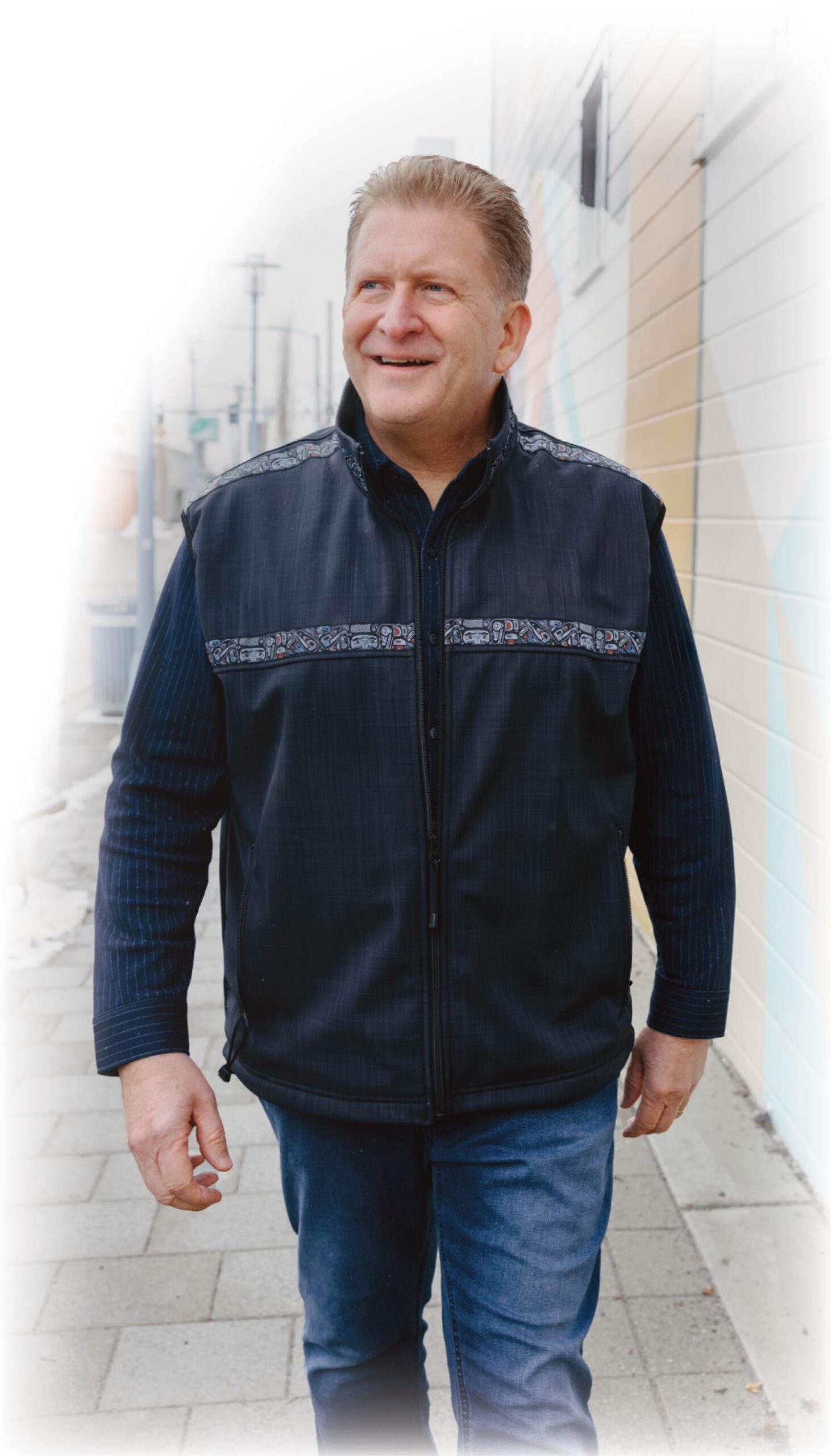 Portrait photograph close-up partial white layered gradient view of Bruce Dingeman smiling in a dark navy blue jacket vest and dark blue denim jeans as he walks outside on a sidewalk somewhere nearby a building as he looks off into the distance