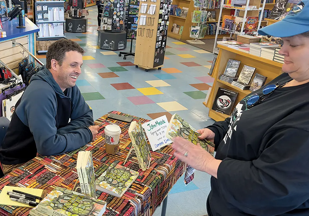 cropped view of John Messick sitting and smiling at an author table in a book store, a woman standing in front of the table surveys the cover of Messicks memoir “Compass Lines: Journeys Toward Home”
