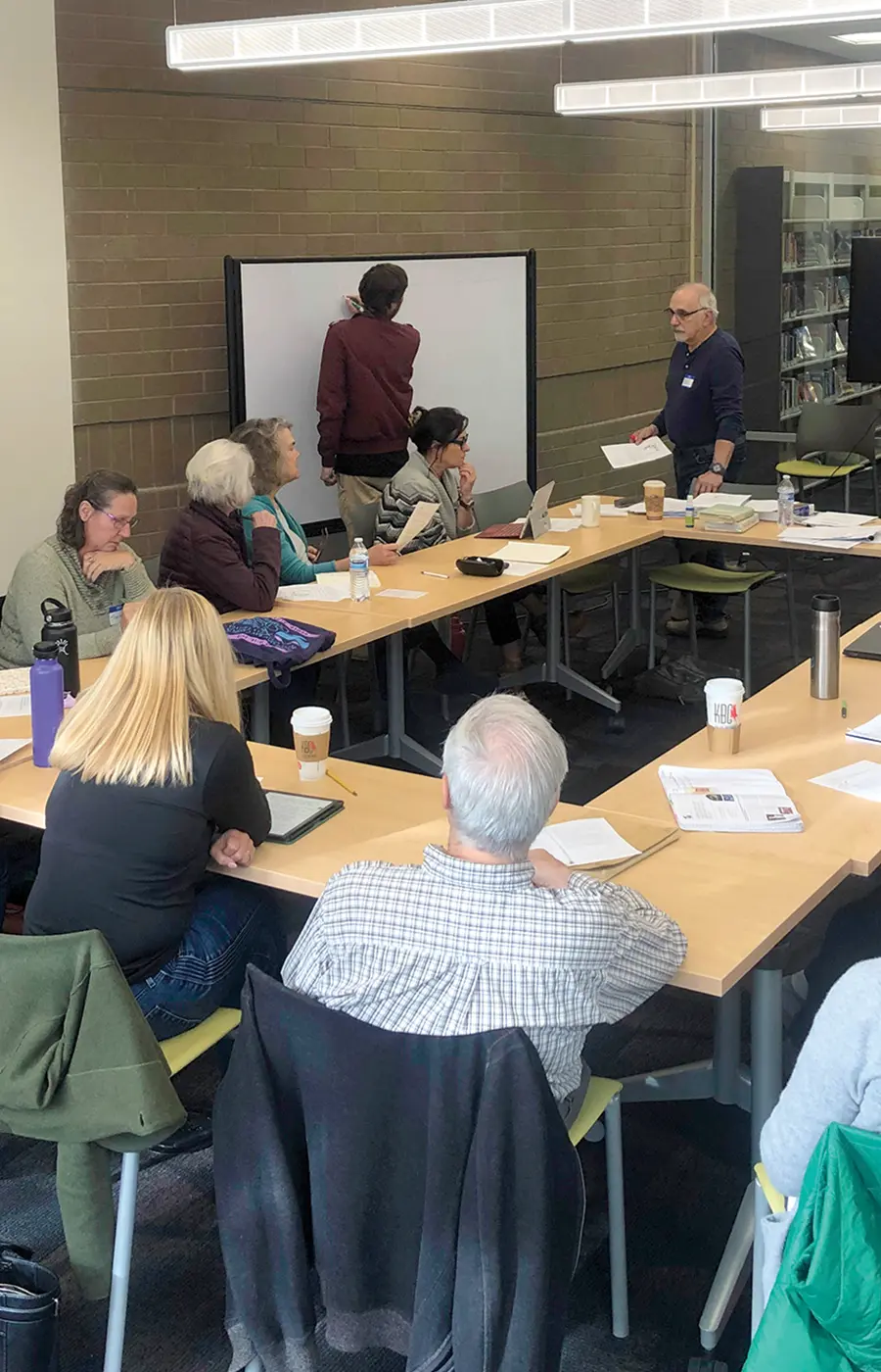 adults of varying ages sit around desks shaped in a rectangle in a small library conference room, a man stands at a whiteboard during an activity session held by 49 Writers