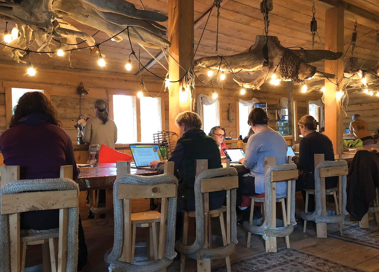 a large cabin like room with a long central table, at the table and other countertops in the room many authors work on laptops in a workshop setting