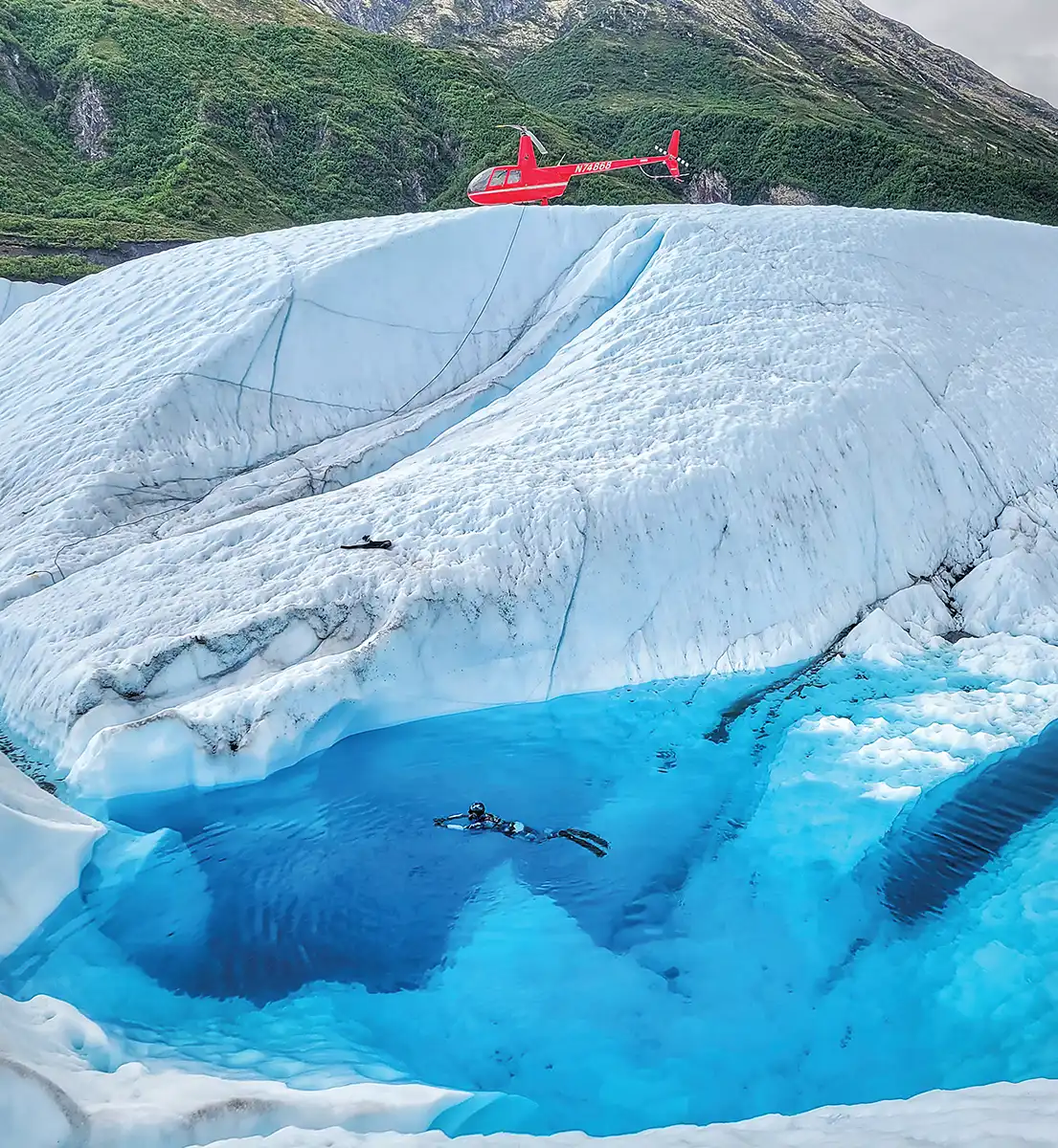 high angle, distant view of a diver swimming in water in a glacial crater, a red helicopter sit on the other edge of the glacier