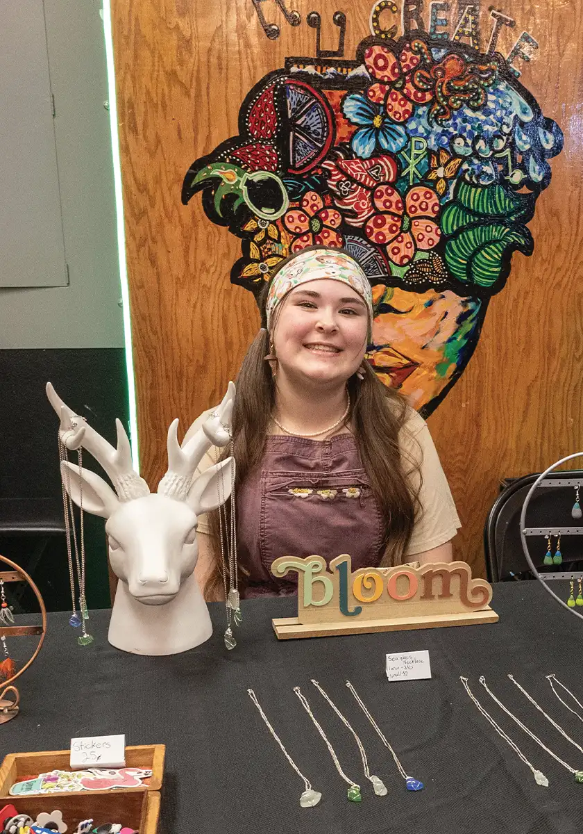 a young proprietress wearing a headband and two ponytails smiles while sitting at her jewelry table at the Kid Business Fair