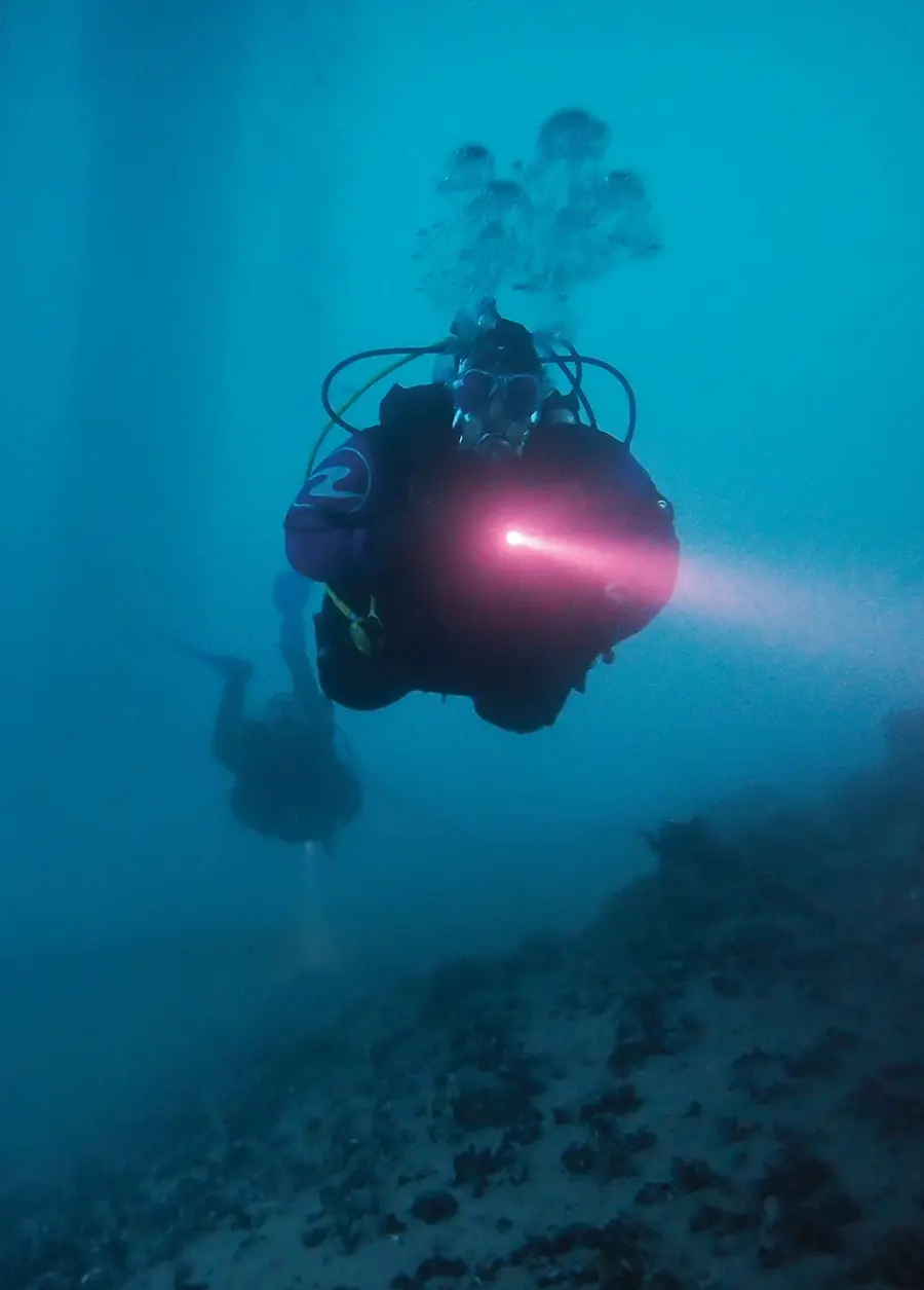 dark view of a diver holding a flashlight, another diver swims out of focus in the background both exploring winter Alaskan waters