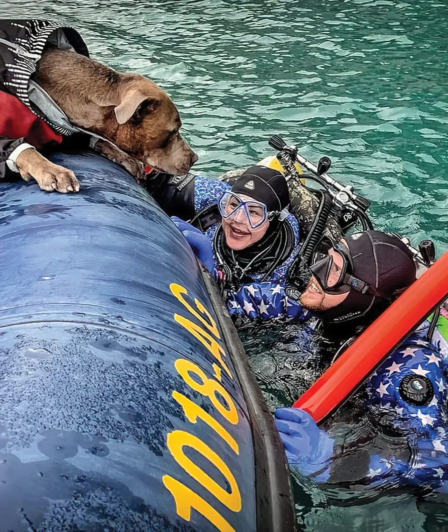 a woman and a man float beside a raft at the surface of a body of water while wearing scuba gear, they smile while looking at Bruce, a brown dog leaning over the side of the raft and looking back at the two divers