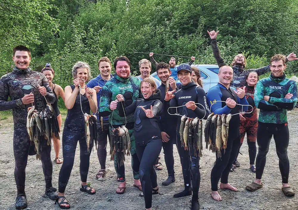 a large group of freediving spearfishers take a shore side photo while in their wet suits, some of the divers present heavy duty fish stringers loaded with invasive Northern pike