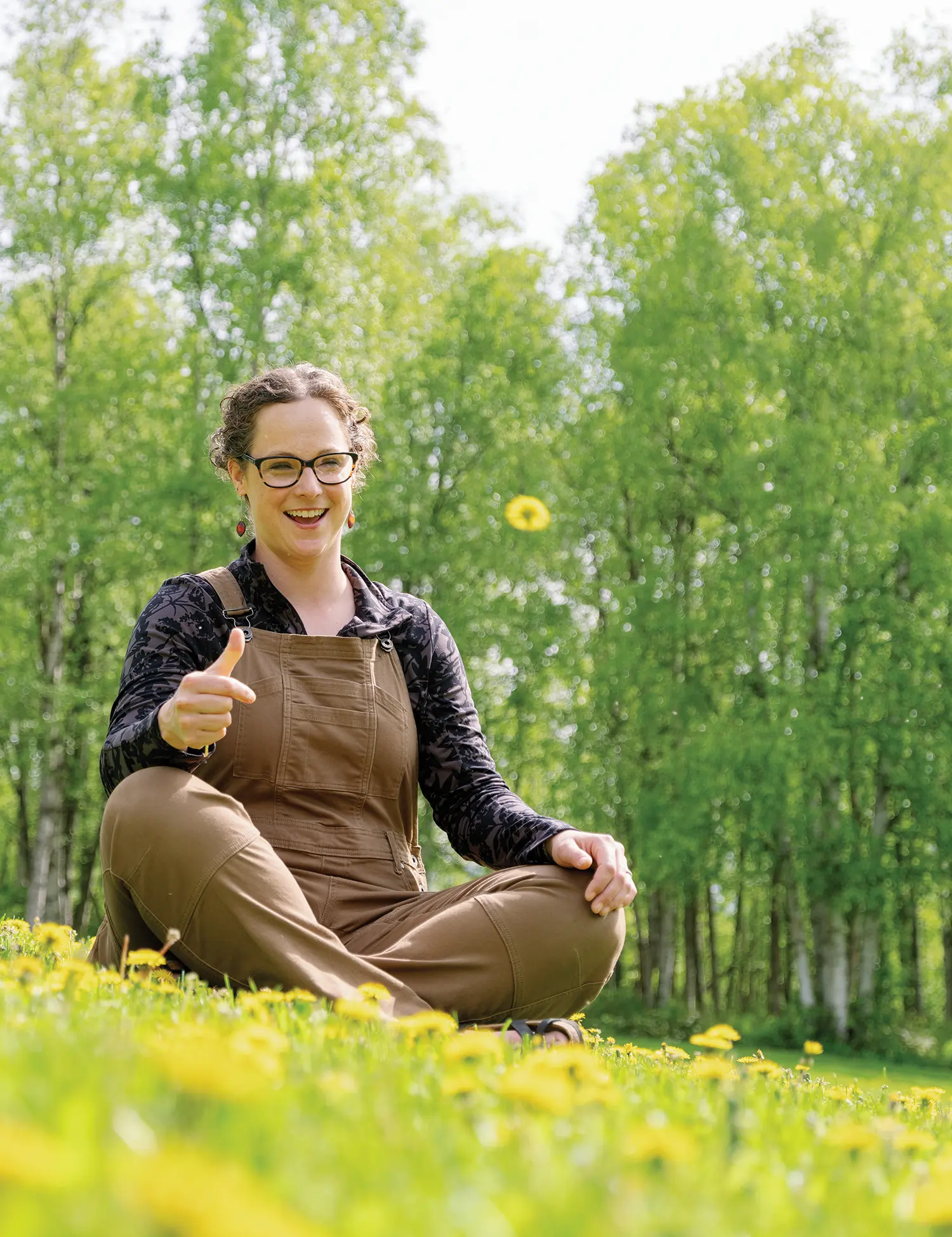 Portrait photograph close-up view of Emily Longbrake smiling at something off into the distance as she is posing with her right hand thumb up in the air while she is seated down in a flowery field grassland setting outside on a clear sunny day with a bunch of green lush trees behind her in the distant background; Emily is wearing a dark brown denim overall dress on top of her dark violet flower-shape pattern long-sleeve shirt, black outer frame see-through prescription glasses, and brown amber colored earrings