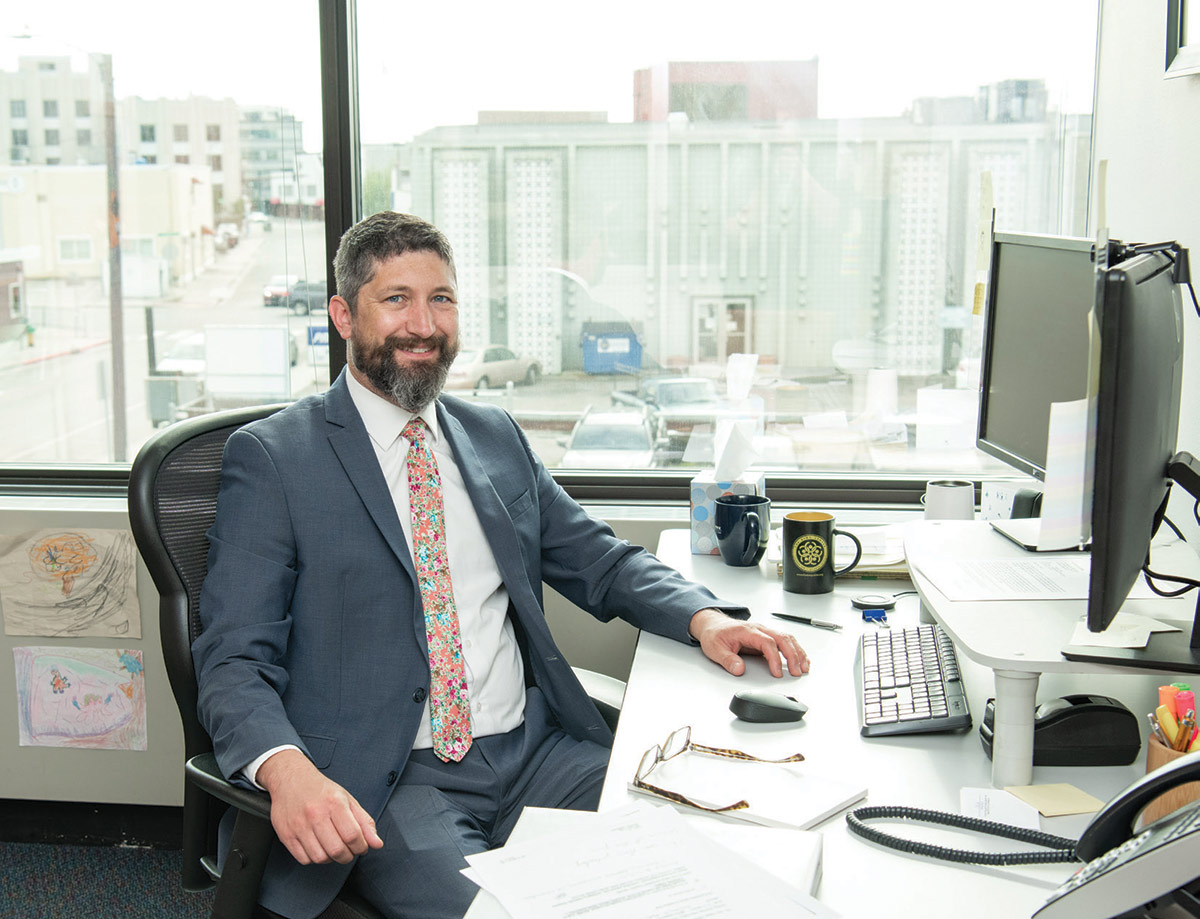 Steven Hansen smiling in his office while sitting at his desk with window and city view in the background