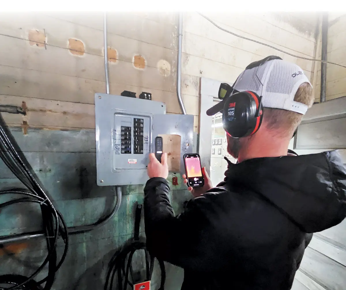 Partial white gradient background covering the top of a close-up portrait photograph view of a man in a white snapback hat with his black sunglasses on top of his hat and black/burnt orange headphones around his ears while he has a black puffy jacket equipped as he uses his smartphone and another different curved thin round skinny shaped electronic device tool to inspect a dark grey open electrical power switch box enclosure attached to the wall of a rusty building area indoors