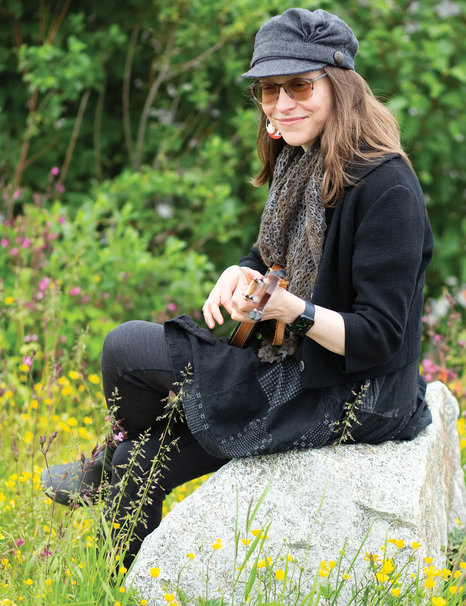 Portrait photograph close-up view of Amy O'Neill Houck grinning in a dark grey rounded shaped flat cap, dark bronze tinted inner frame see through sunglasses, a smart watch equipped on her left wrist, a black button-up long-sleeve cardigan dress equipped with a dark brown/dark marine blue scarf around her neck, faded black denim jeans, white/red/black custom circular shaped cultural earrings equipped, and black dress boots equipped as she holds a dark brown small ukulele instrument in her hands as she is seated on top of a light grey granite rock outside in a flower field area