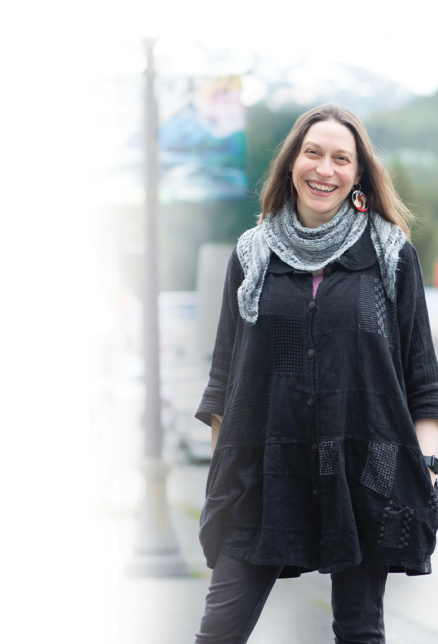 Mostly white to transparent gradient background covering the portrait photograph close-up view of Amy O'Neill Houck smiling as she is standing somewhere outside in a town sidewalk area on a mostly cloudy, overcast day; Amy is wearing a black button-up long-sleeve cardigan dress with a light grey scarf around her neck and white/red/black custom circular shaped cultural earrings; She also has on a smart watch on her left wrist plus has her hands inside the pockets of the cardigan dress as she has on faded black denim jeans equipped