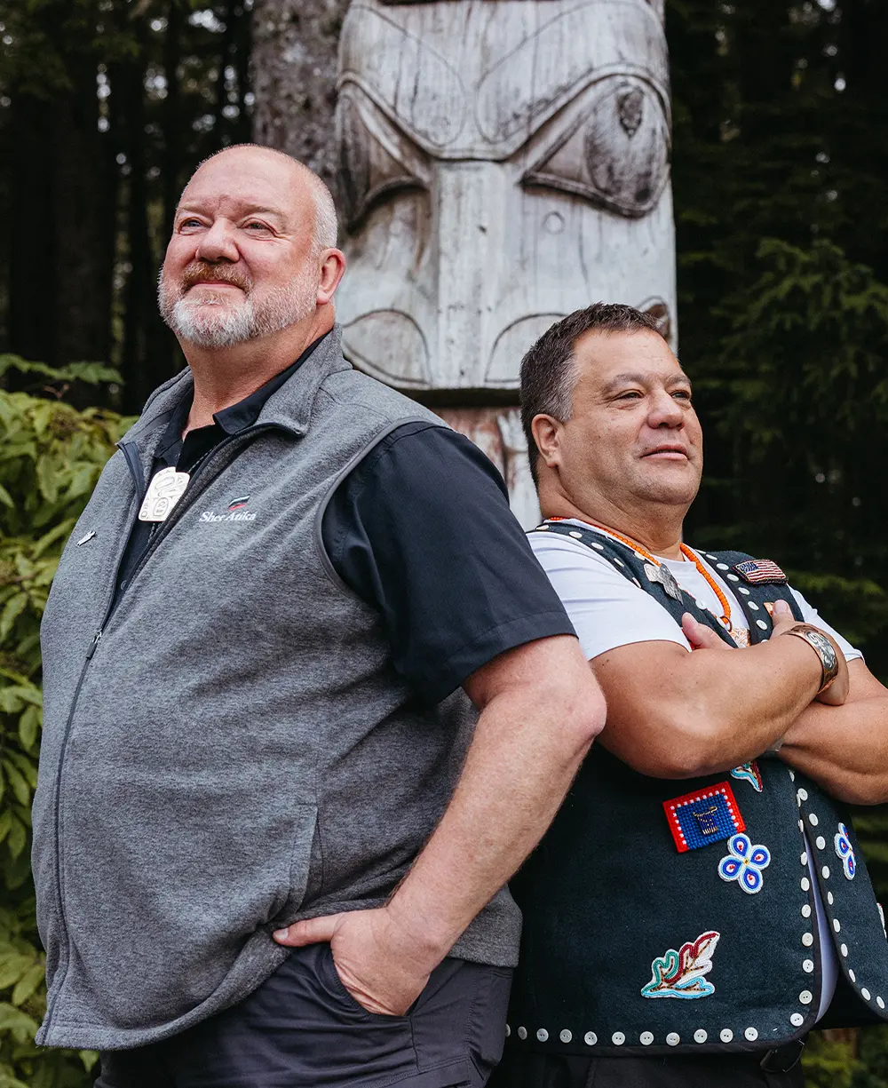 Shee AtikÁ chair Steve Karpstein and vice chair Larry Garrity stand almost back to back in front of a totem pole