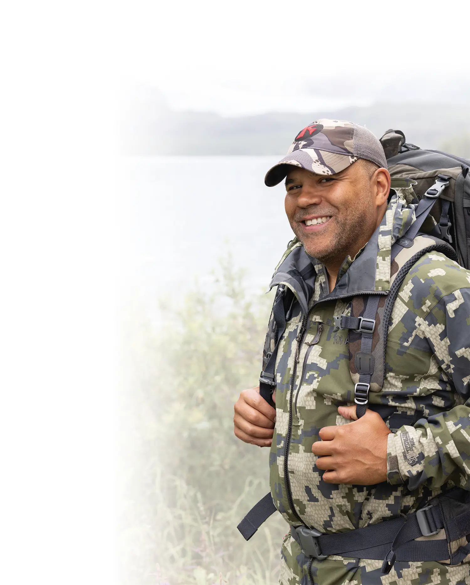 Mostly white gradient background covering the portrait photograph close-up view of Russell Dick smiling as he is standing somewhere outside in some lush greenery bushes nearby a body of water on a mostly cloudy, overcast day; Russell is wearing a dark forest green, light tan/beige, and dark grey mixed camoflauge jacket with a dark grey hiking backpack equipped behind on his back and a light tan/beige plus dark grey mixed camoflauge hat