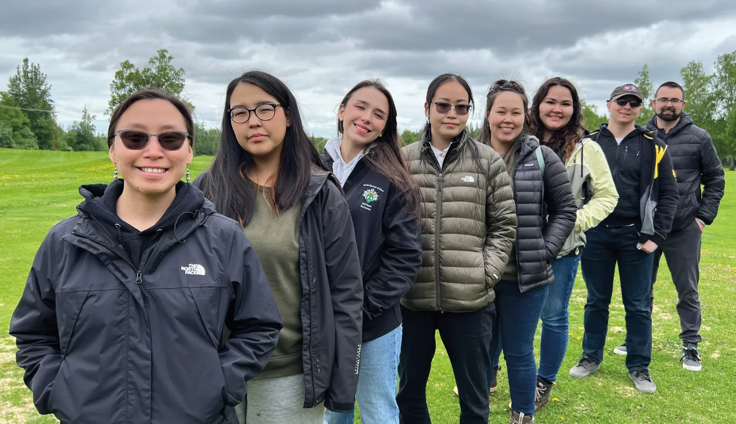 A group of nine people stands in a line outdoors, all dressed in jackets, posing for a photo on a grassy field under a cloudy sky.