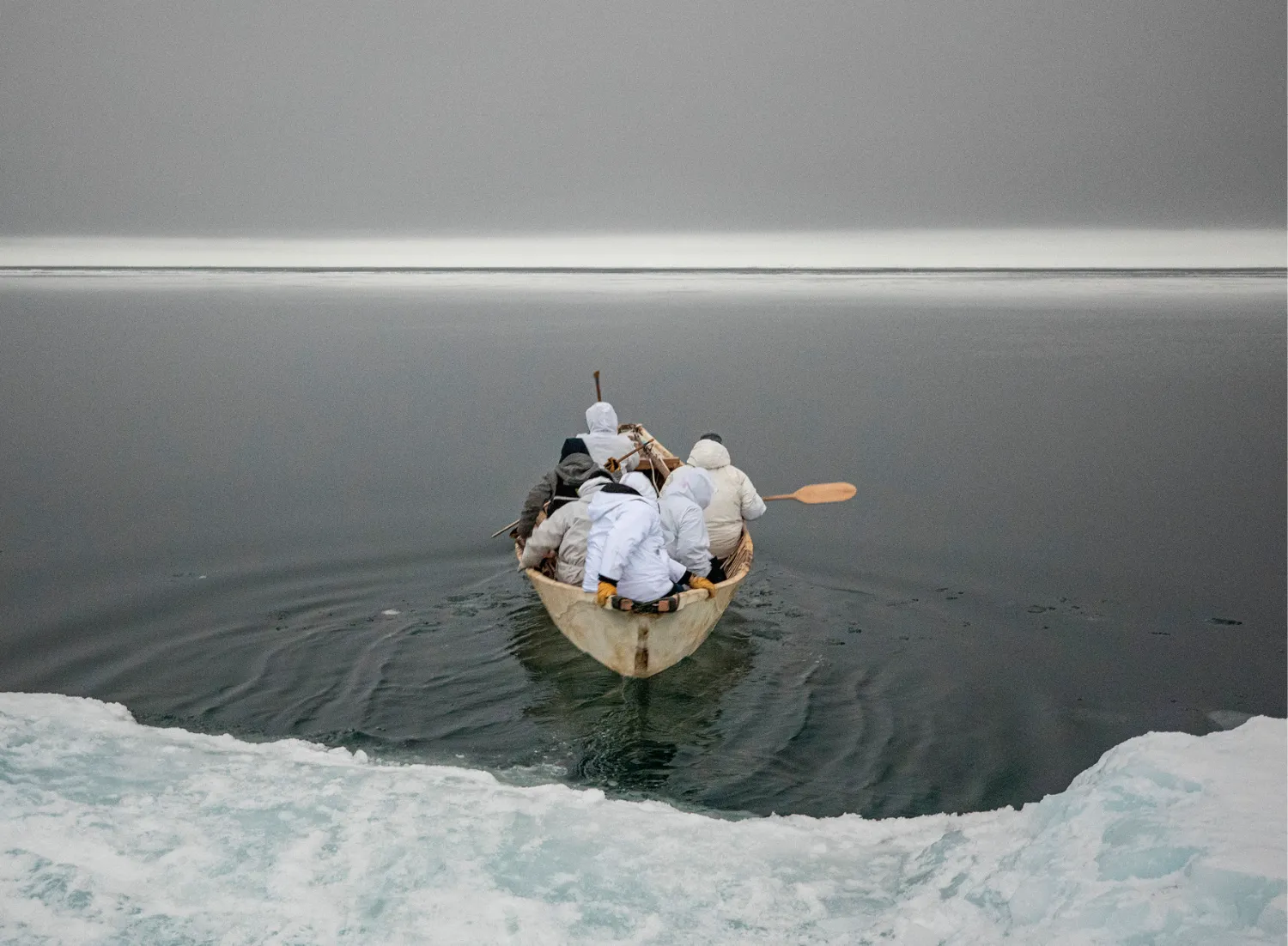 A group of people in white hooded jackets paddle a small wooden boat through dark waters surrounded by ice, with a foggy horizon in the distance.