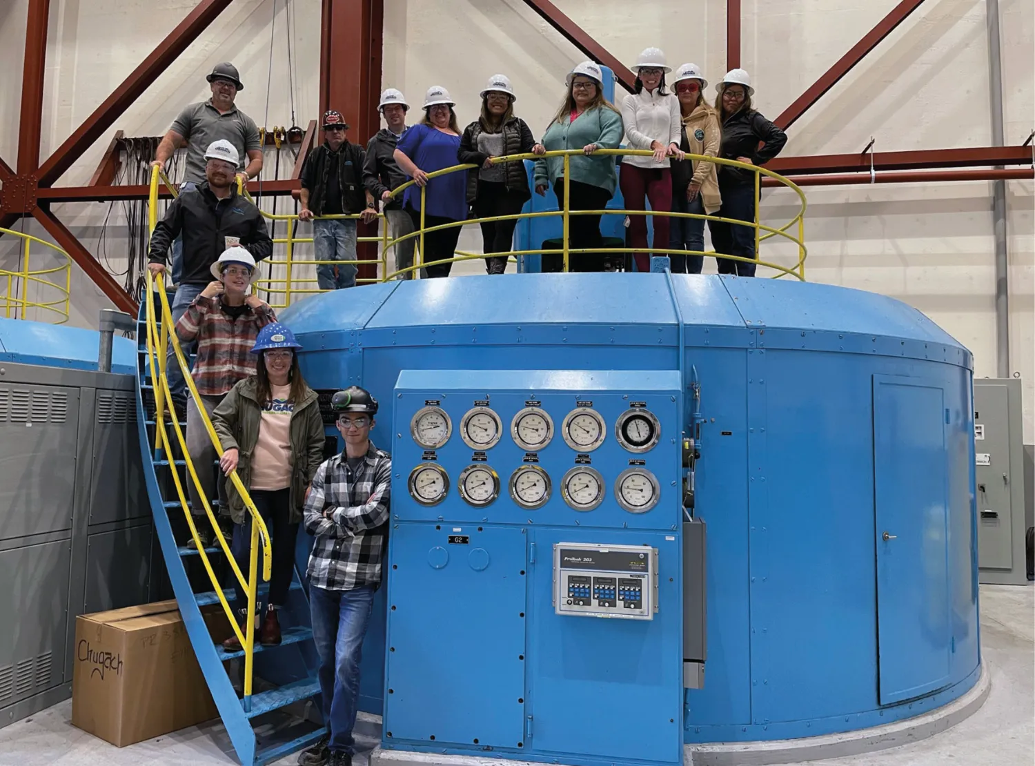 A group of people wearing hard hats pose on and around a large blue industrial machine, standing on metal platforms in a factory setting.