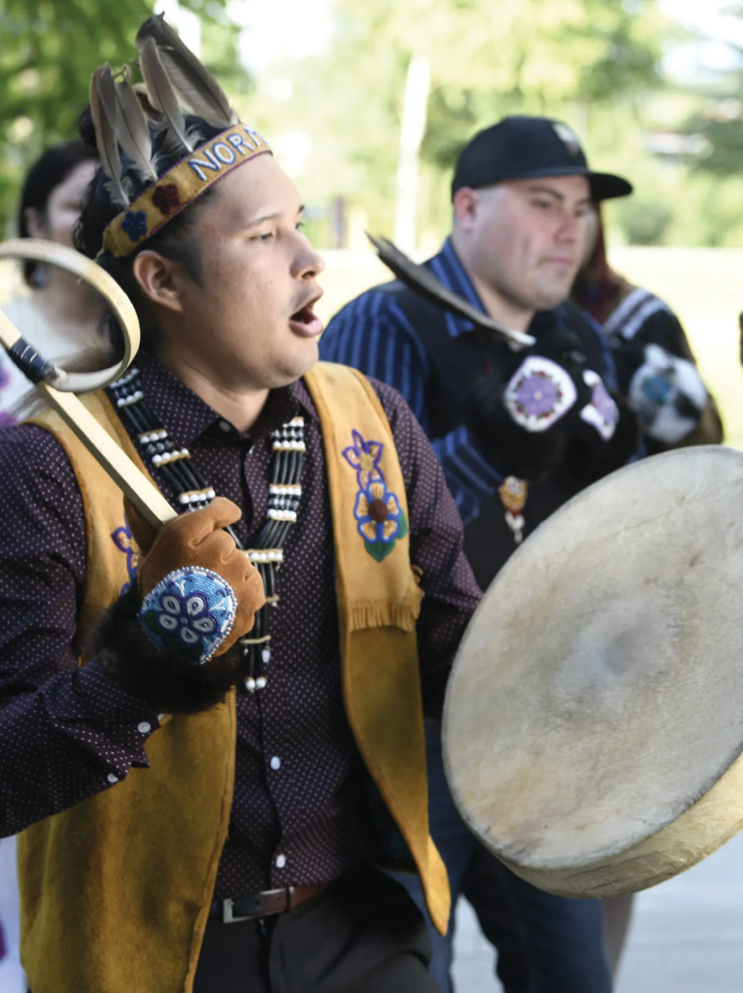 A man wearing a feathered headdress and traditional regalia sings and plays a drum, participating in a cultural performance outdoors.