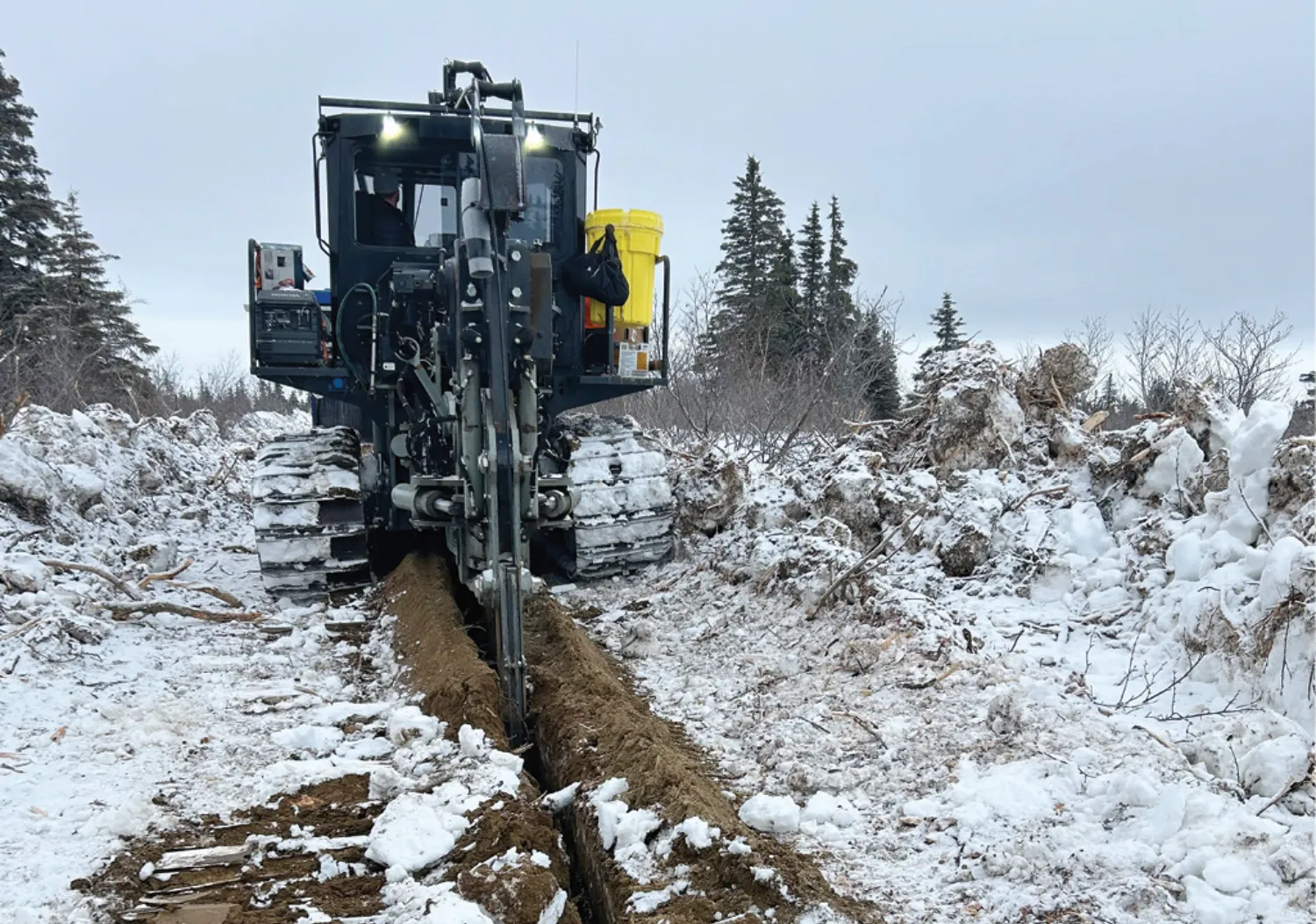 A heavy machine with large tracks digs a trench through snowy terrain, surrounded by trees and snow-covered piles of dirt.